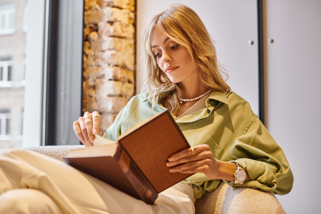 Una mujer sentada en un sofá absorbida en un libro en un acogedor apartamento. - Foto, imagen
