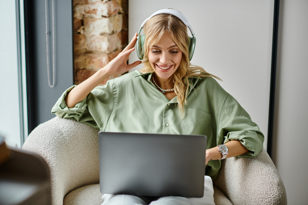 Kobieta siedząca w domu na krześle, używająca laptopa i nosząca słuchawki. - Zdjęcie, obraz