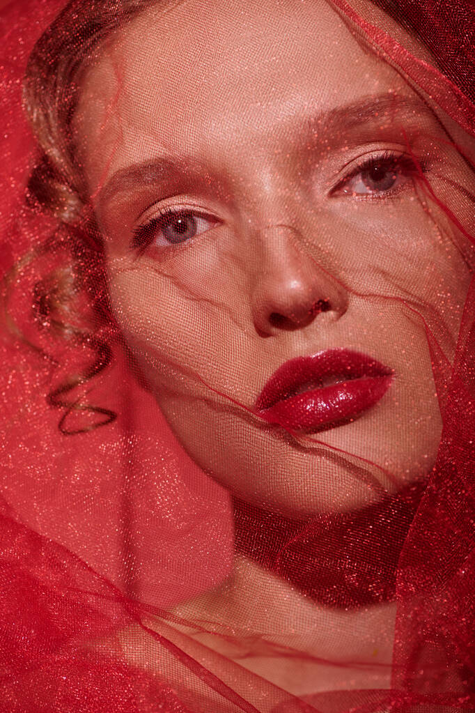 Eine junge Frau strahlt klassische Schönheit aus, als sie mit einem roten Schleier posiert, der ihren Kopf in einem Studio-Setting schmückt. - Foto, Bild
