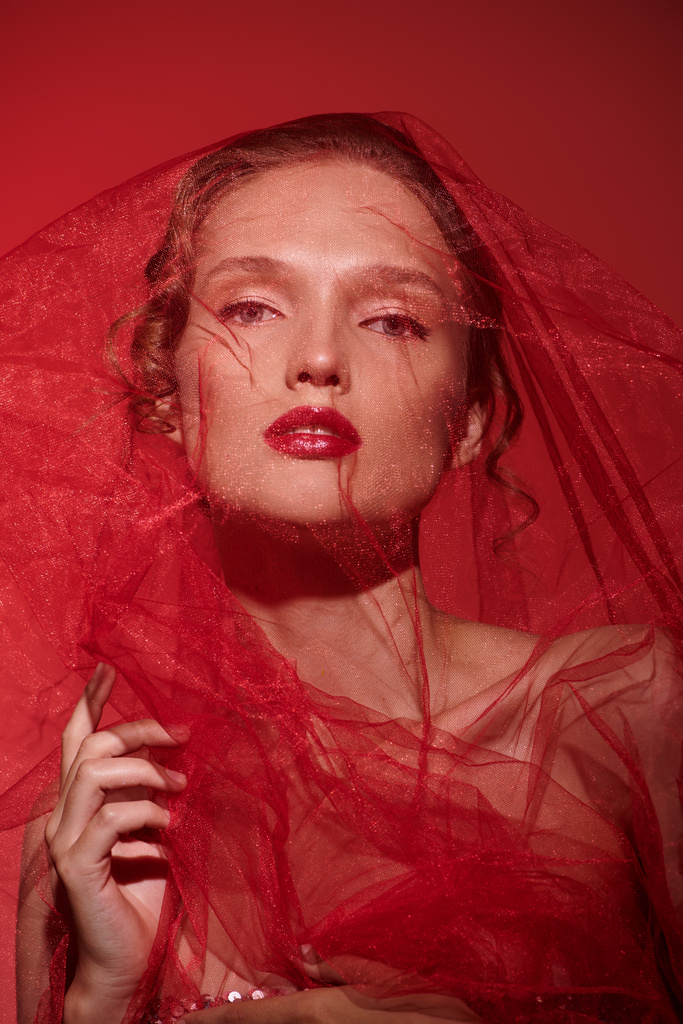 Μια νεαρή γυναίκα αποπνέει κλασική ομορφιά ενώ ποζάρει σε ένα στούντιο με ένα εντυπωσιακό κόκκινο φόρεμα με ένα πέπλο που απλώνεται πάνω από το κεφάλι της.. - Φωτογραφία, εικόνα