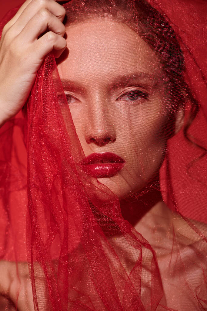Uma jovem exala beleza clássica, seu cabelo ruivo em cascata sob um impressionante véu vermelho em um estúdio contra um fundo preto. - Foto, Imagem