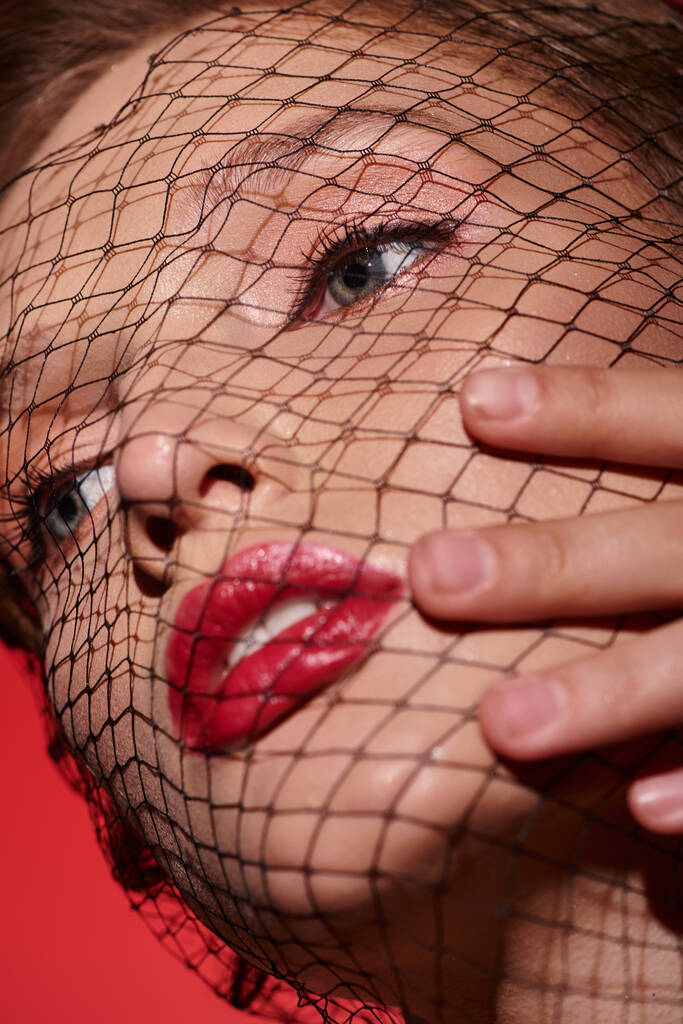 Tajemnicza kobieta z czerwoną szminką i siatką zakrywającą twarz emanuje klasycznym pięknem i intrygą w studio. - Zdjęcie, obraz