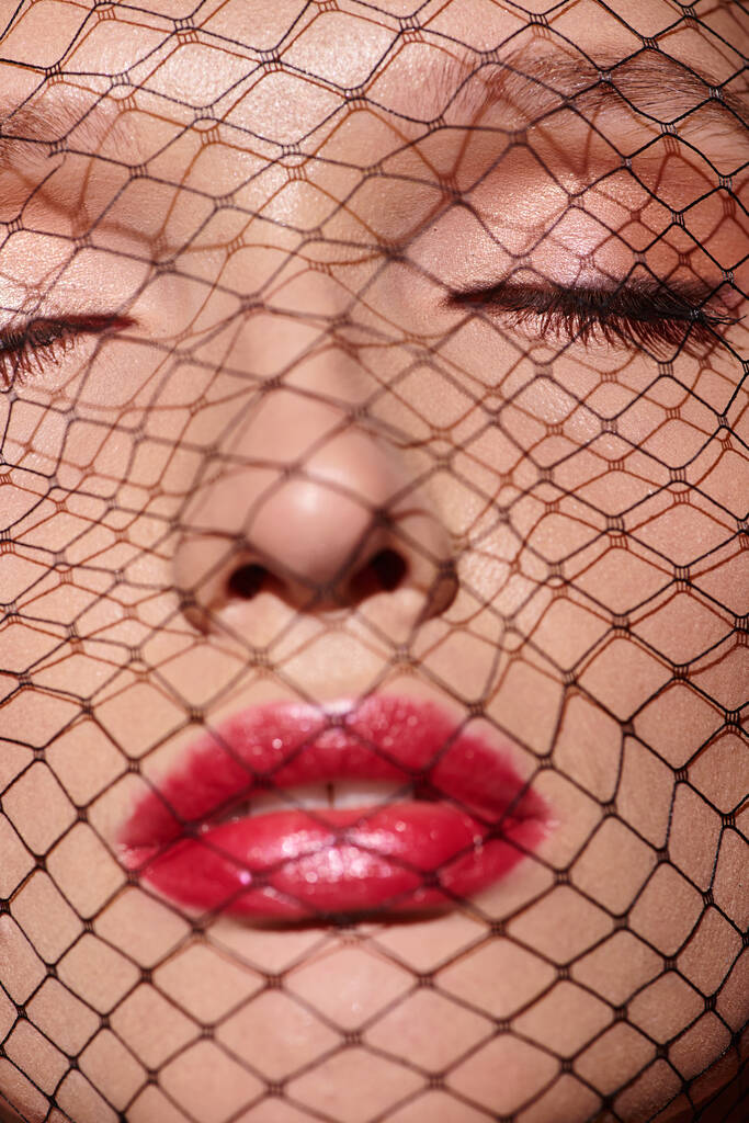 Een klassieke schoonheid met rode lippenstift staart door een net dat haar gezicht bedekt in een mystieke en betoverende pose. - Foto, afbeelding