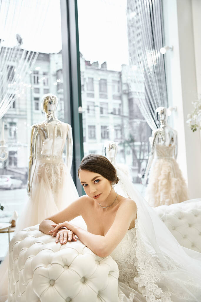 Eine junge brünette Braut im Brautkleid sitzt auf einer Bank vor einem Fenster. - Foto, Bild