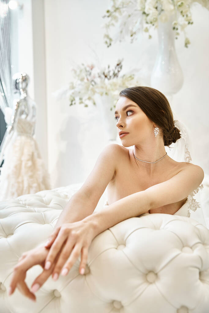 Mladá brunetka nevěsta ve svých svatebních šatech sedí půvabně na bílém lůžku, obklopená aurou klidu a krásy. - Fotografie, Obrázek