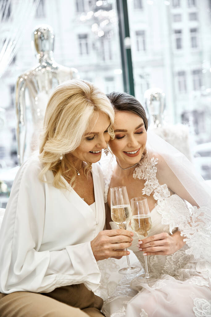 Νεαρή νύφη με νυφικό φόρεμα και η μητέρα της κάθονται μαζί, κρατώντας ποτήρια κρασιού στο σαλόνι νυφικών. - Φωτογραφία, εικόνα