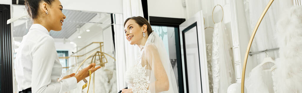 Egy fiatal barna menyasszony menyasszonyi ruhában, és a bevásárló asszisztense a tükör előtt áll egy menyasszonyi szalonban.. - Fotó, kép