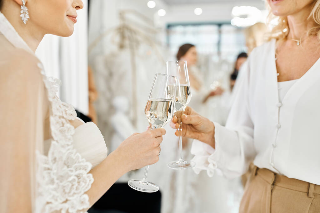Eine junge brünette Braut im Hochzeitskleid steht mit ihrer Mutter mittleren Alters, beide halten Champagnergläser in einem Brautsalon. - Foto, Bild