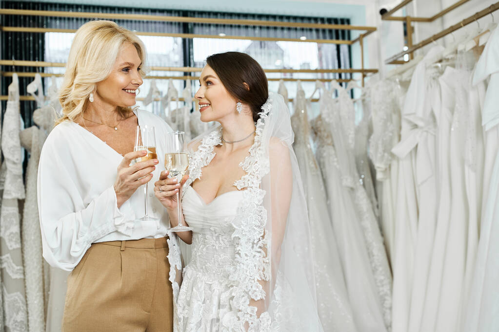 結婚式のドレスとブライダルサロンで一緒に立っている彼女の中年の母親の若いブルネットの花嫁. - 写真・画像