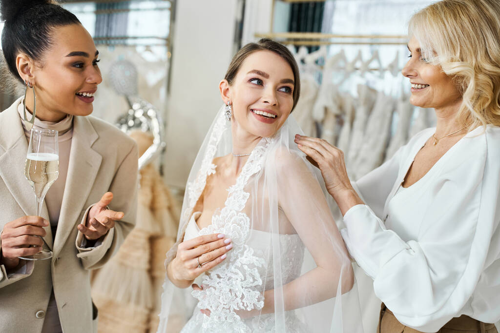 Een jonge brunette bruid in een trouwjurk glimlacht opgewonden terwijl haar moeder en beste vriendin van middelbare leeftijd, een bruidsmeisje, haar helpen een sluier aan te trekken. - Foto, afbeelding