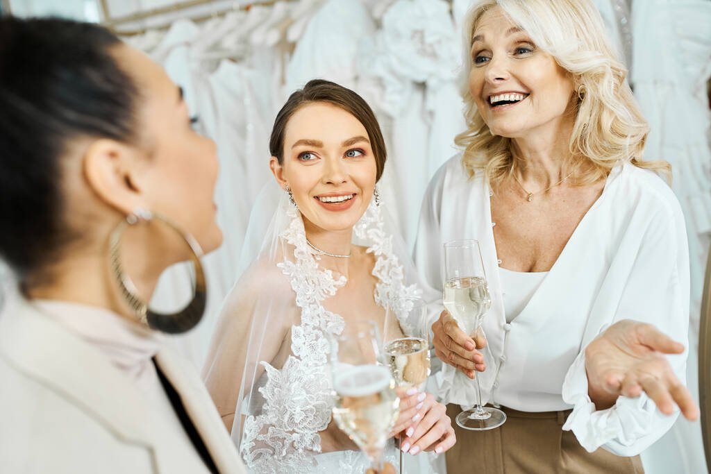 Una joven novia con un vestido de novia, su madre de mediana edad, y una dama de honor en un salón de novias, sosteniendo copas de vino. - Foto, imagen