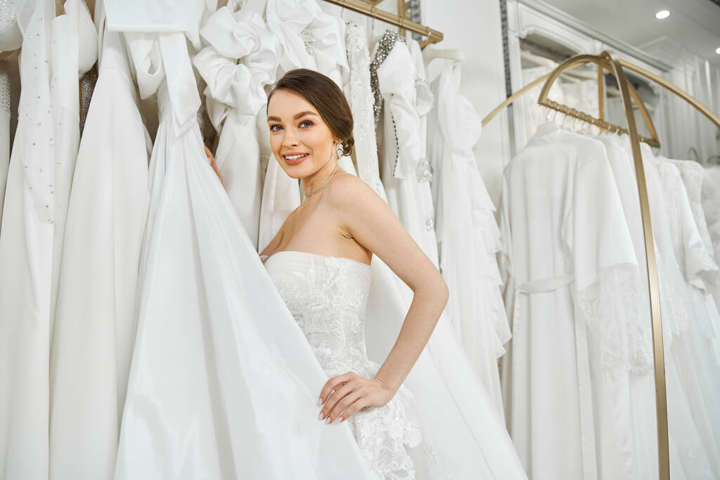 Una joven novia morena se para frente a un estante de vestidos blancos, eligiendo cuidadosamente su vestido de novia perfecto. - Foto, imagen