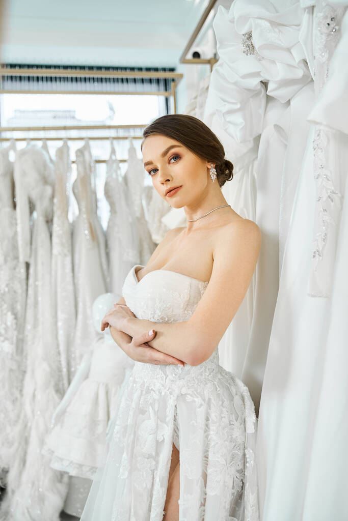 Eine junge brünette Braut steht zwischen einem Kleiderständer in einem Hochzeitssalon und wählt ihr perfektes Kleid. - Foto, Bild