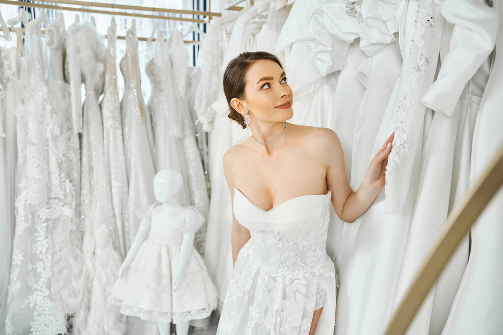 Mladá, krásná brunetka stojící před regálem šatů ve svatebním salónu, vybírající si perfektní šaty. - Fotografie, Obrázek