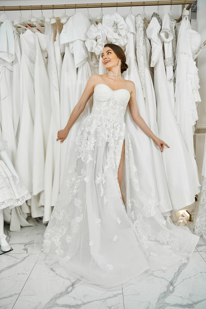 Μια νέα, όμορφη νύφη θαυμάζει ένα ράφι φορέματα σε ένα σαλόνι γάμου, μελετώντας το τέλειο φόρεμα της. - Φωτογραφία, εικόνα