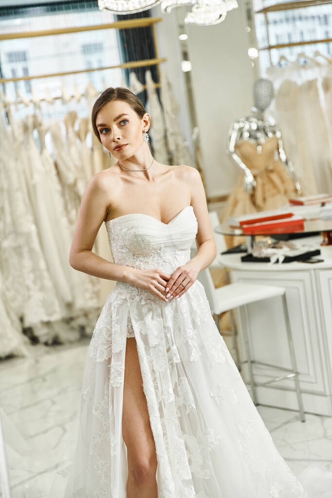 Fiatal barna menyasszony egy fehér ruha bámulja a tükör egy esküvői szalonban, megtestesítő elegancia és a kegyelem. - Fotó, kép
