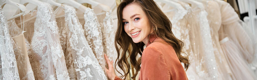 Una joven novia se pone delante de una selección de vestidos de novia, tratando de encontrar el vestido perfecto para su día especial. - Foto, Imagen