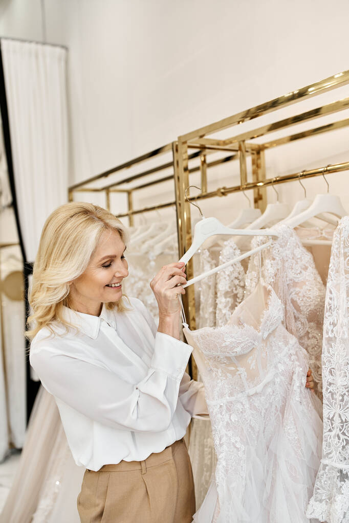 Eine schöne Verkäuferin mittleren Alters hilft einer Frau in einem Salon beim Stöbern in Hochzeitskleidern auf einem Regal. - Foto, Bild