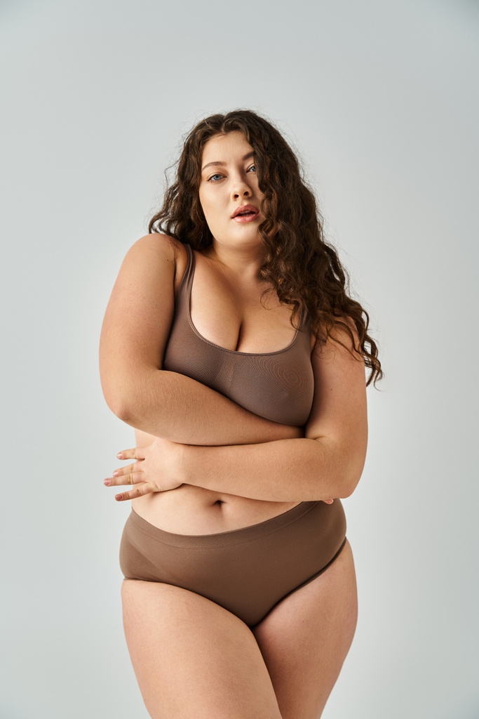 alluring plus size young woman in underwear with curly brown hair hugging herself on grey background - Photo, Image