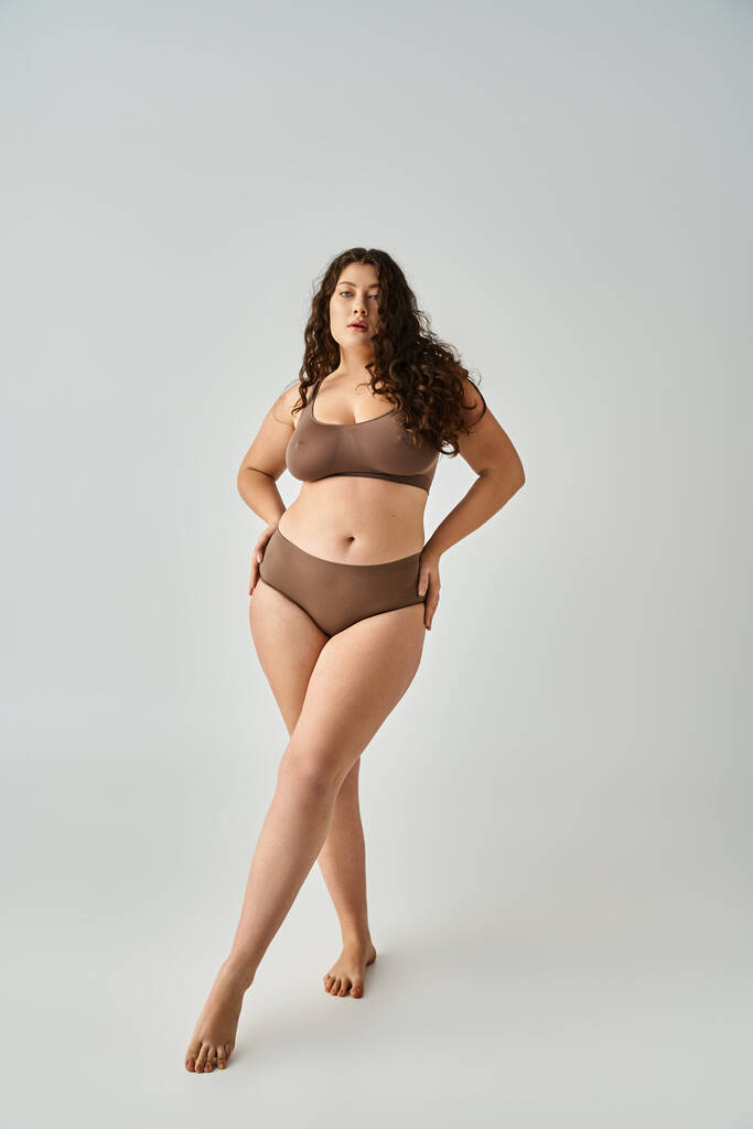 beautiful plus size young woman in underwear with hands on hips and cross legs in grey background - Photo, Image