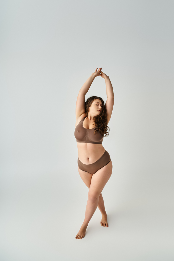 charming plus size girl in underwear with closed eyes putting hands to up in grey background - Photo, Image