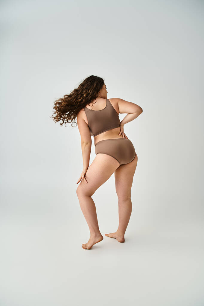 plus size young woman in underwear with curly brown hair posing from back on grey background - Photo, Image