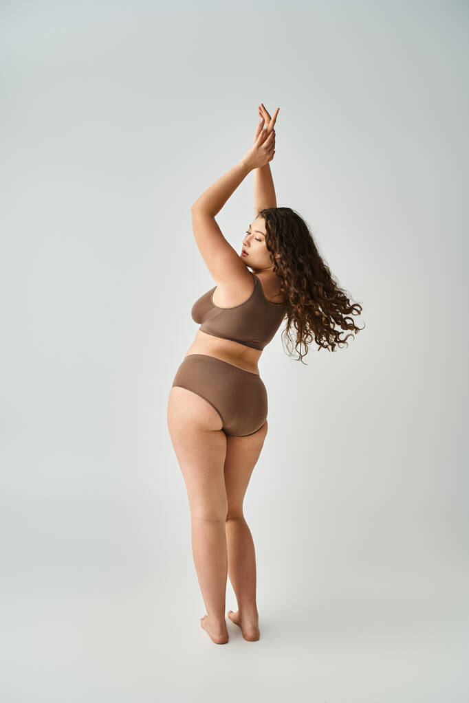 séduisante femme courbée en sous-vêtements avec les cheveux bruns bouclés posant du dos et mettant les mains vers le haut - Photo, image