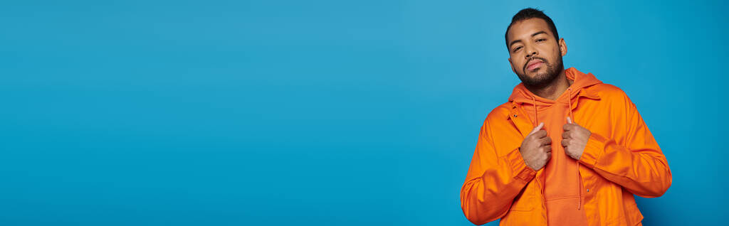 Mavi arka planda, turuncu elbiseli çekici Afrikalı Amerikalı genç adamın yatay görüntüsü. - Fotoğraf, Görsel