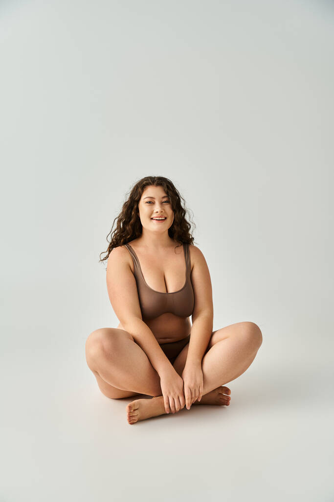 cheerful curvy young woman in brown underwear sitting cross legged against grey background - Photo, Image
