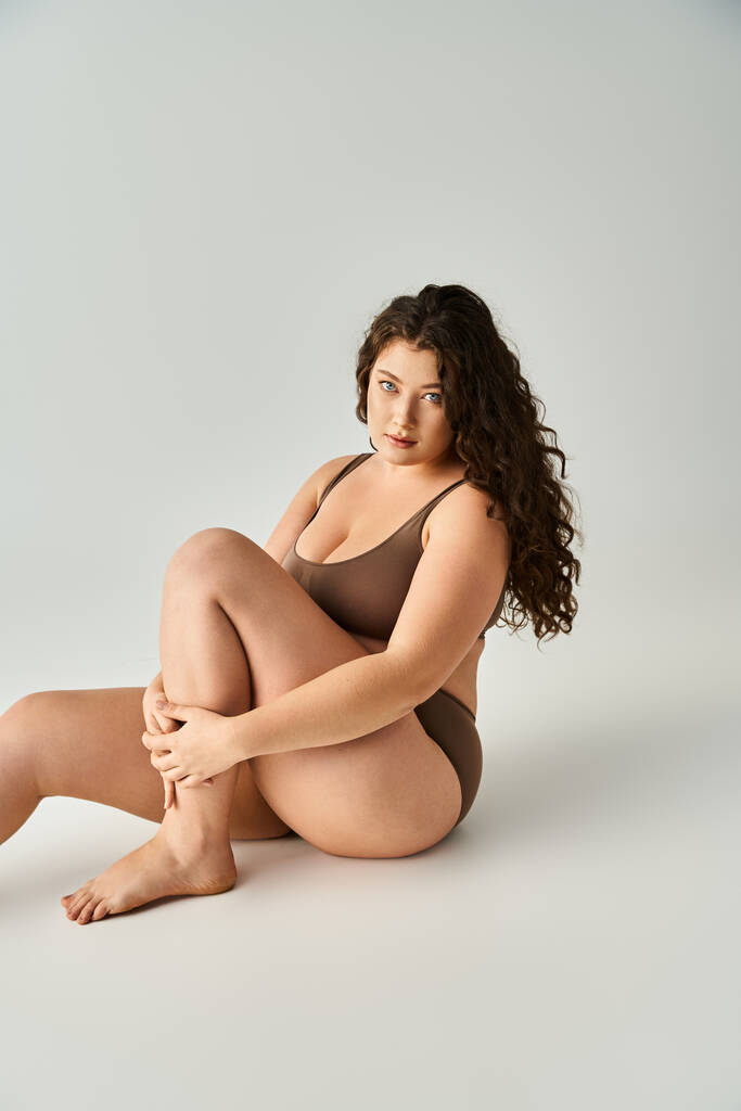 aantrekkelijke plus size jonge vrouw in bruin ondergoed zit en knuffelen been met armen - Foto, afbeelding