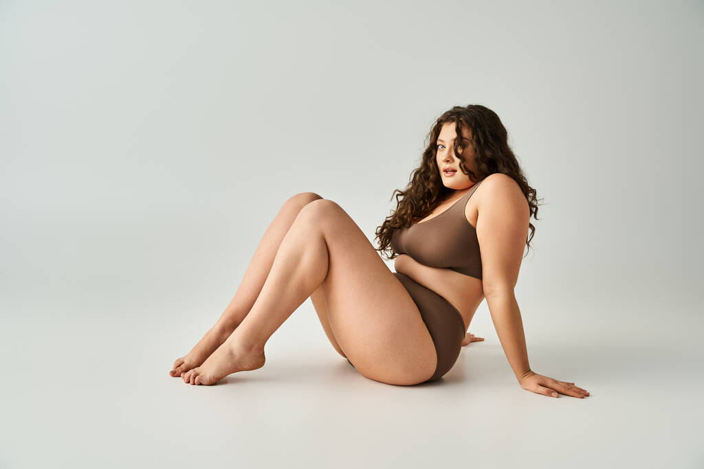 приваблива жінка плюс розмір в коричневій білизні сидить, спираючись на руки і тягнучи ноги до неї - Фото, зображення