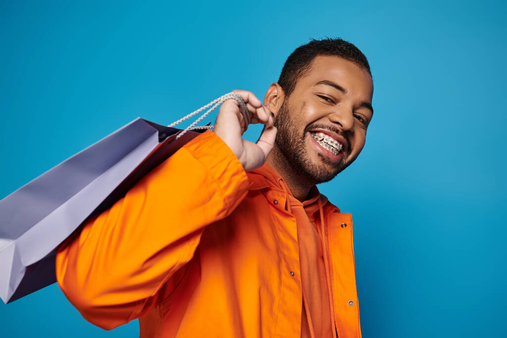 усміхнений афроамериканський чоловік в помаранчевому вбранні з торговою сумкою над плечем на синьому фоні - Фото, зображення