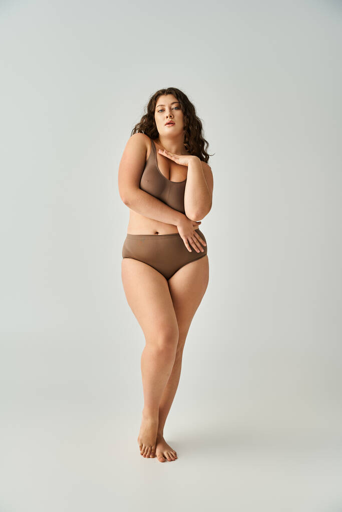 seductive plus size woman in brown lingerie with curly hair hugging herself on grey background - Photo, Image