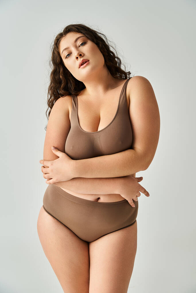 alluring plus size young girl in brown lingerie with curly hair hugging herself on grey background - Photo, Image