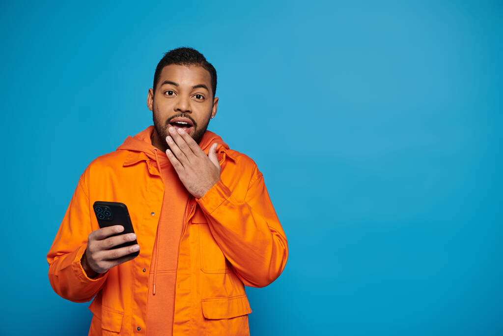 surprised african american man in orange outfit scrolling in social media and covering hand to mouth - Photo, Image