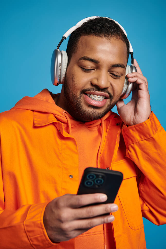 gelukkig Afrikaans amerikaanse man in oranje outfit en koptelefoon luisteren naar muziek met smartphone - Foto, afbeelding