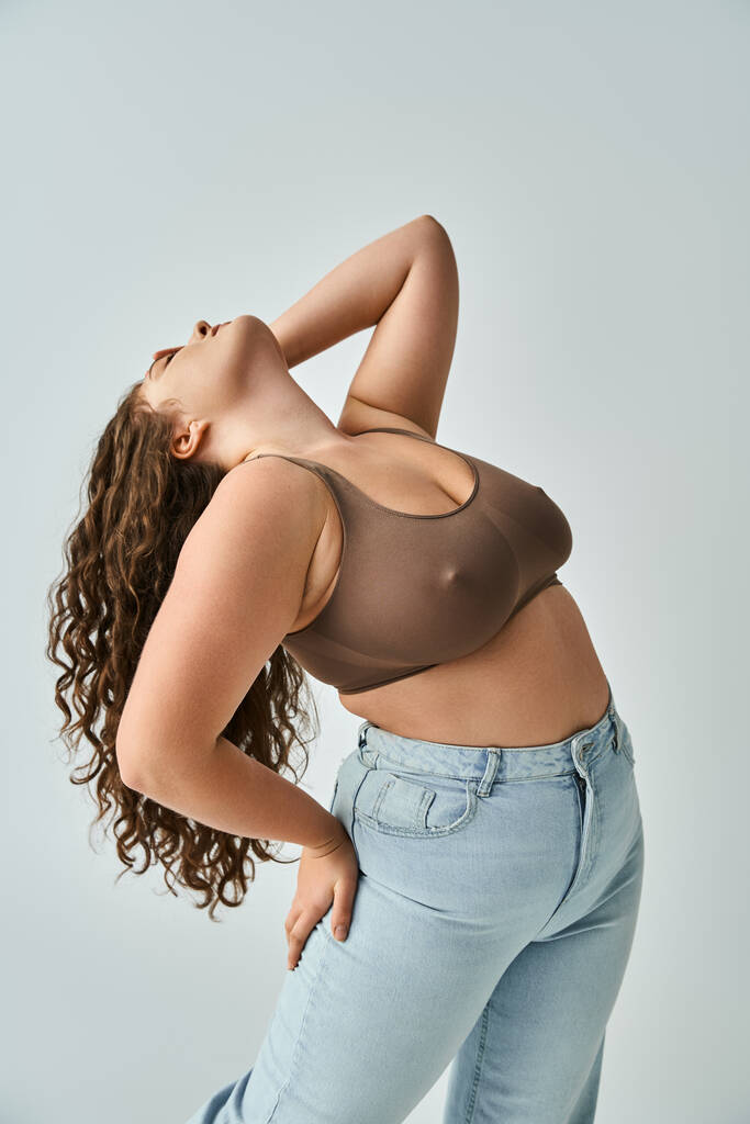plus size young woman in brown bra and blue jeans with curly hair putting head to behind with hand - Photo, Image