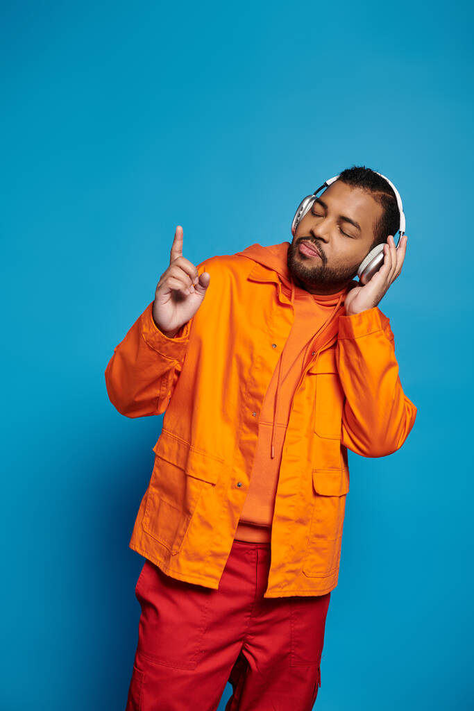 usmívající se africký Američan v oranžové oblečení a sluchátka poslech hudby na modrém pozadí - Fotografie, Obrázek