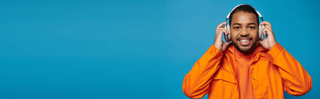 Banner eines afrikanisch-amerikanischen Mannes in orangefarbenem Outfit blickt in die Kamera und hält mit den Händen Kopfhörer - Foto, Bild