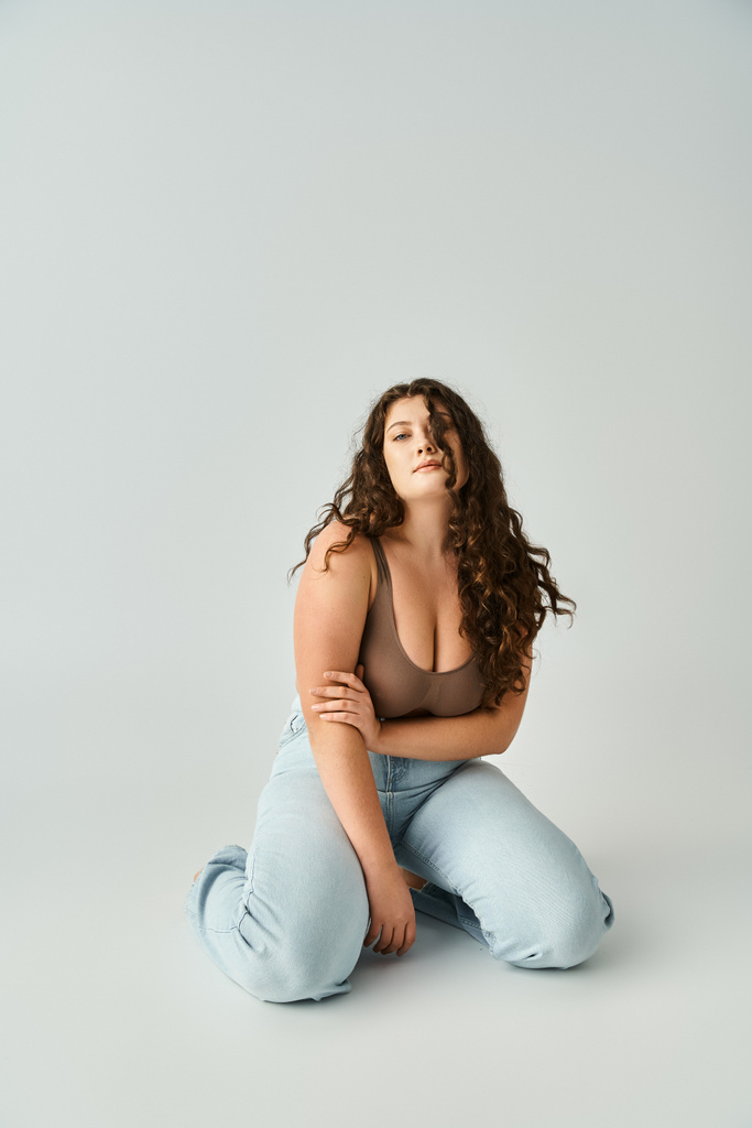 alluring plus size young woman in brown bra and blue jeans sitting and touching with hand to arms - Photo, Image