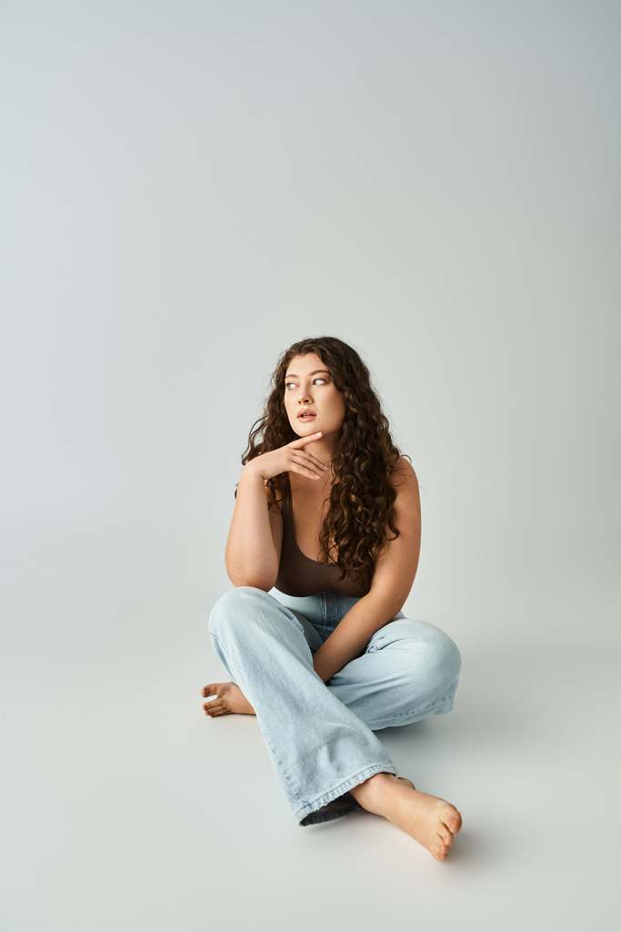 atraente plus tamanho jovem menina em sutiã marrom e jeans azul sentado e tocando o dedo para queixo - Foto, Imagem