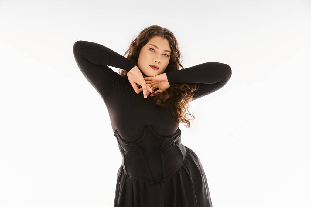 seductora chica joven de tamaño grande en traje elegante negro con el pelo rizado posando con las manos en gris - Foto, imagen