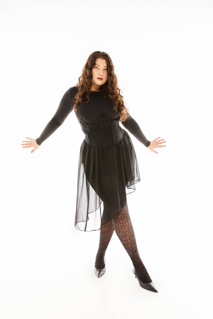glamourous καμπυλωτή νεαρή γυναίκα σε μαύρο κομψό ντύσιμο με τα χέρια στο πλάι και σταυρωτά πόδια σε γκρι - Φωτογραφία, εικόνα