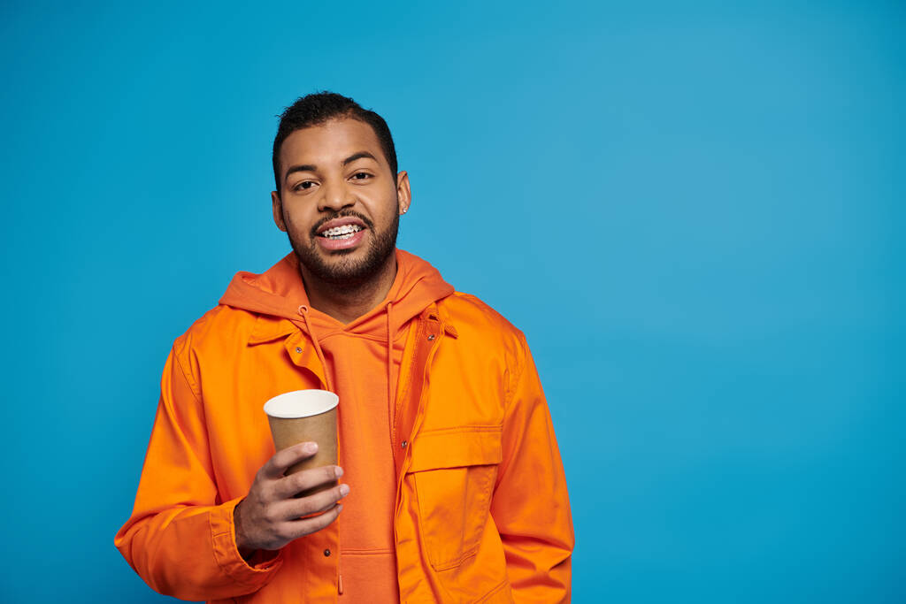 20代の陽気なアフリカ系アメリカ人男性は,青い背景に対して紙コップを手に - 写真・画像