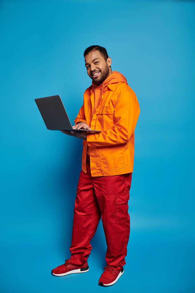 glimlachende Afro-Amerikaanse man in oranje outfit staan zijdelings met laptop in handen - Foto, afbeelding