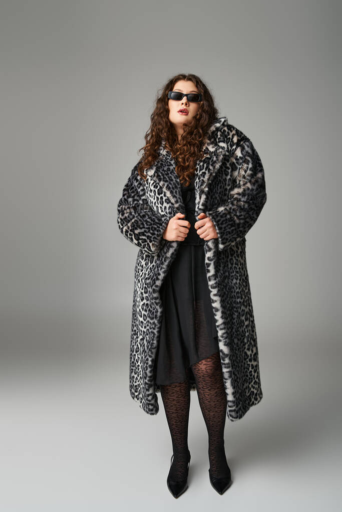 mooie ronde vrouw in luipaard bont jas en zonnebril staan op grijze achtergrond - Foto, afbeelding