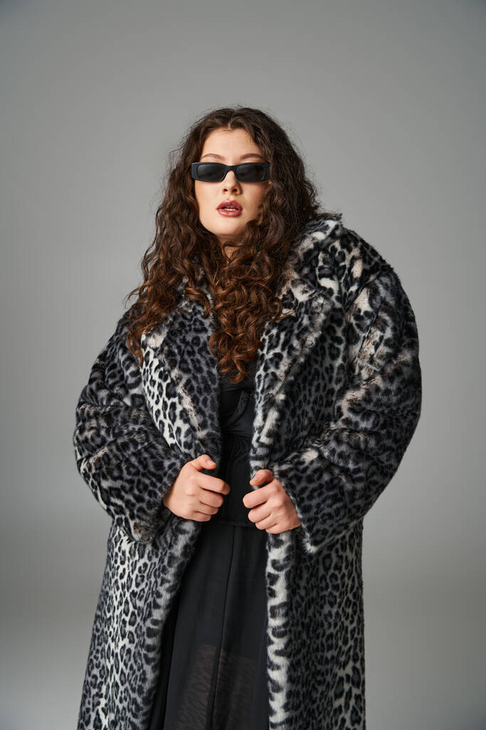 ελκυστική συν μέγεθος νεαρή γυναίκα σε λεοπάρδαλη γούνα παλτό και γυαλιά ηλίου στέκεται σε γκρι φόντο - Φωτογραφία, εικόνα