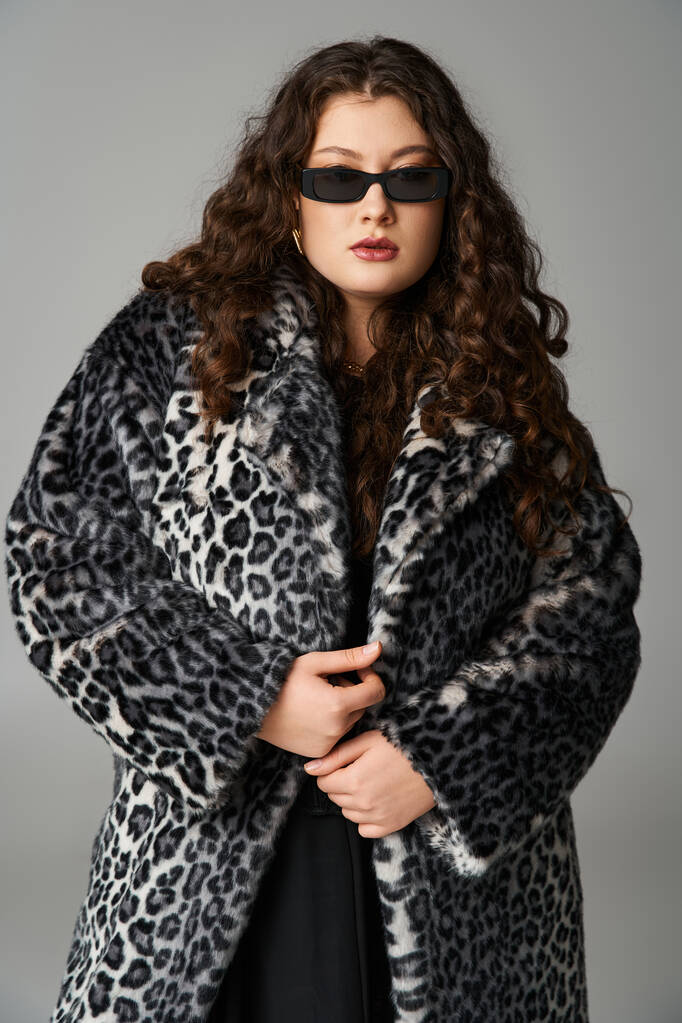 mujer joven elegante más tamaño en abrigo de piel de leopardo y gafas de sol de pie sobre fondo gris - Foto, Imagen