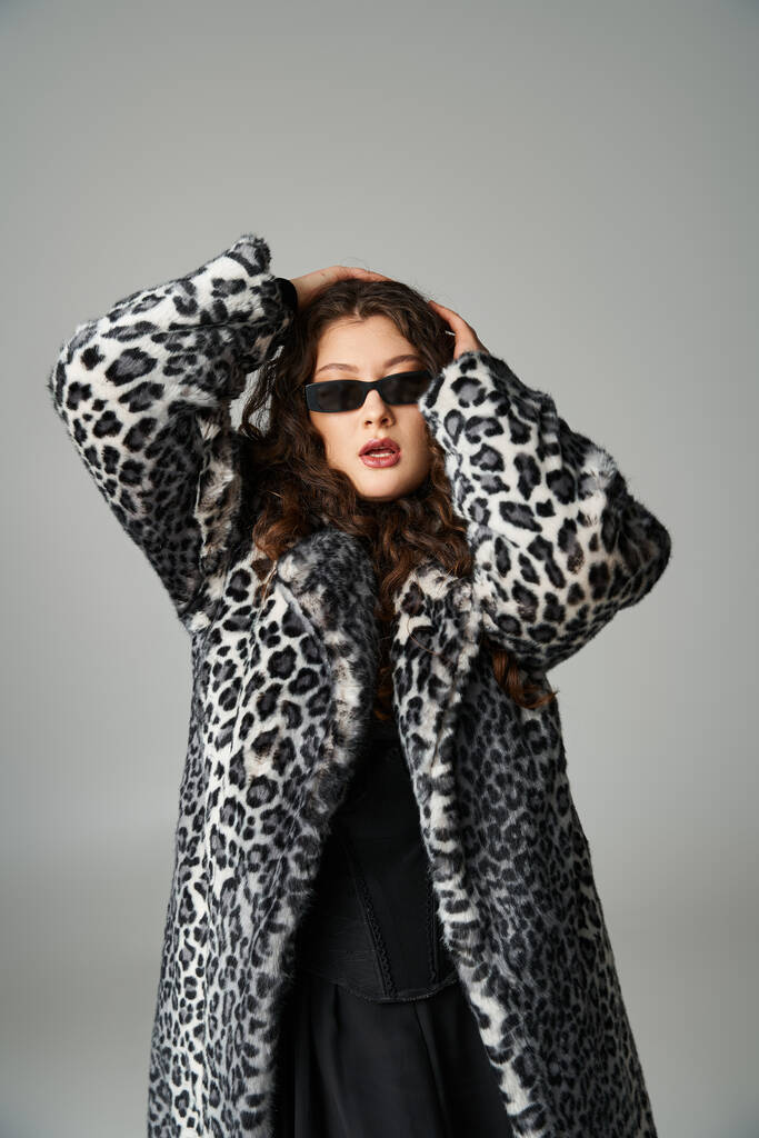 hermosa mujer con curvas en abrigo de piel de leopardo y gafas de sol abrazando la cabeza con las manos sobre fondo gris - Foto, imagen