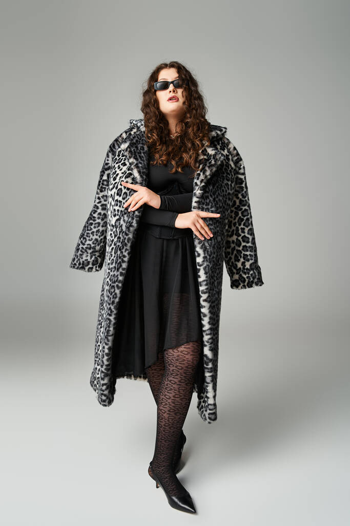 seductora mujer con curvas en gafas de sol de pie con abrigo de piel de leopardo sobre fondo gris - Foto, Imagen